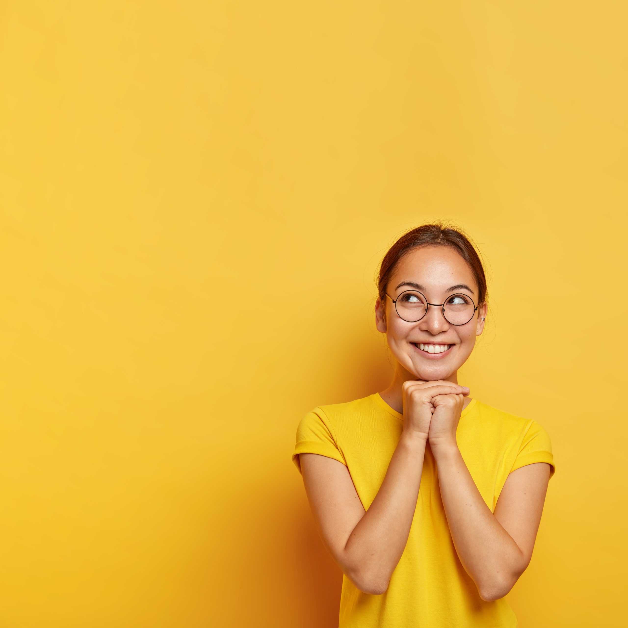 Vertical shot of cheerful thoughtful Asian female keeps hands pressed together under chin, hopes for something awesome, wears spectacles and t shirt, has natural beauty, isolated on yellow studio wall